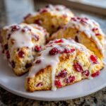 Close-up of several flaky cranberry scones topped with a thick white glaze, served on a white plate.