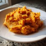 A close-up of fluffy, bright orange mashed sweet potatoes served on a simple white plate.