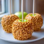 Three homemade caramel apple treats covered in chopped nuts, ready to eat, with a green apple visible in the background.