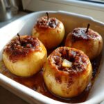 Four golden baked apple treats topped with brown sugar and spices sitting in a white dish.
