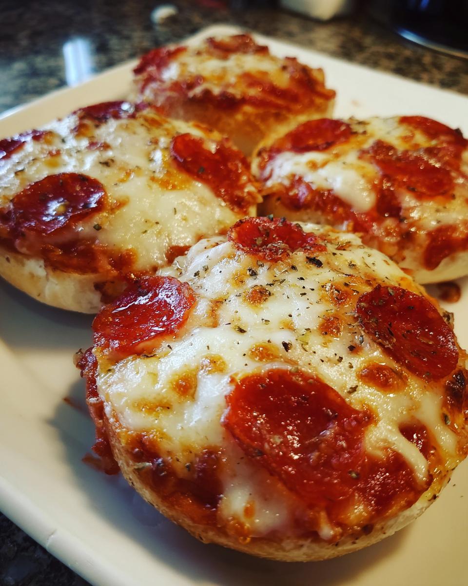 Close-up of four freshly baked pepperoni pizza bagels topped with melted cheese and herbs on a white plate.