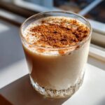 A close-up of a creamy glass of coquito topped generously with ground cinnamon, sitting in bright sunlight.