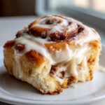 A close-up of a fluffy, warm cinnamon bun generously topped with white cream cheese frosting.