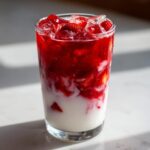 A tall glass showing layered strawberry milk with white milk at the bottom and vibrant red strawberry puree and ice cubes on top.