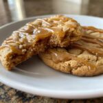 Two soft maple brown sugar cookies on a white plate, one broken open, drizzled with thick caramel sauce.