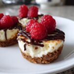 Close-up of a mini cheesecake bite topped with fresh raspberries and chocolate drizzle.
