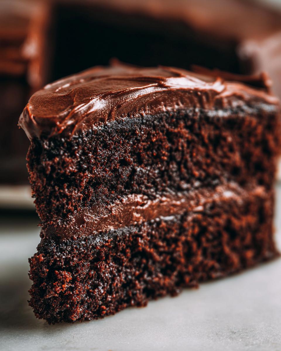 Close-up of a rich, dark slice of chocolate cake recipe with thick, glossy chocolate frosting.