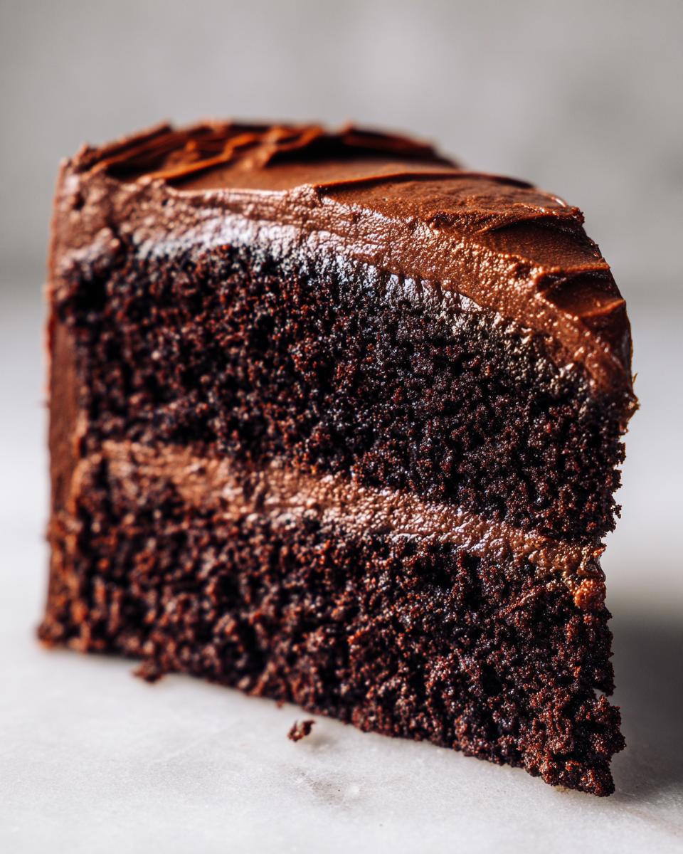 Close-up of a moist, dark slice of chocolate cake recipe layered with thick chocolate frosting.