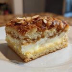 A square slice of bee sting cake featuring layers of sponge, cream filling, and a caramelized almond topping.
