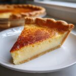 A perfect slice of creamy custard pie with a golden-brown, slightly scorched top, resting on a white plate.