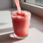 Close-up of a thick, icy watermelon smoothie being poured into a clear glass on a bright white surface.