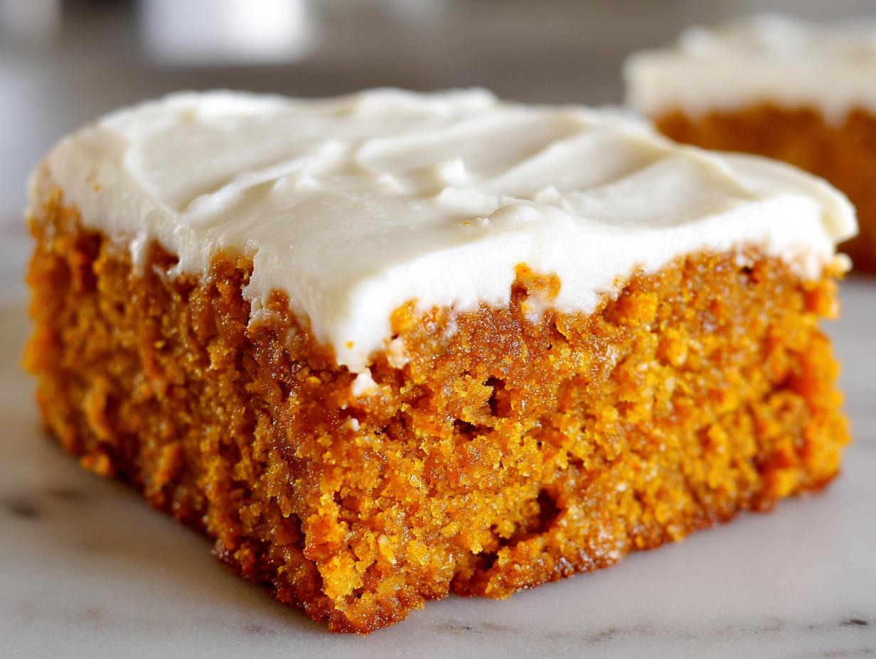 A close-up, side view of a moist, orange pumpkin bar topped with thick, white cream cheese frosting.