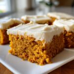 A close-up of a moist square of pumpkin bars with thick cream cheese frosting on a white plate.