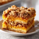 Two stacked squares of rich, orange pumpkin crunch dessert bars with a thick, crumbly topping on a white plate.