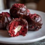 A close-up of several chocolate-dipped red velvet cake balls, one cut open showing the cream cheese center.