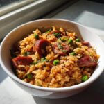 Close-up of a white bowl filled with seasoned sausage and rice mixed with green peas and fresh herbs.