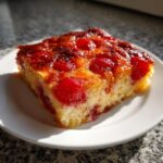 Close-up of a square slice of moist cherry dump cake with a caramelized cherry topping on a white plate.