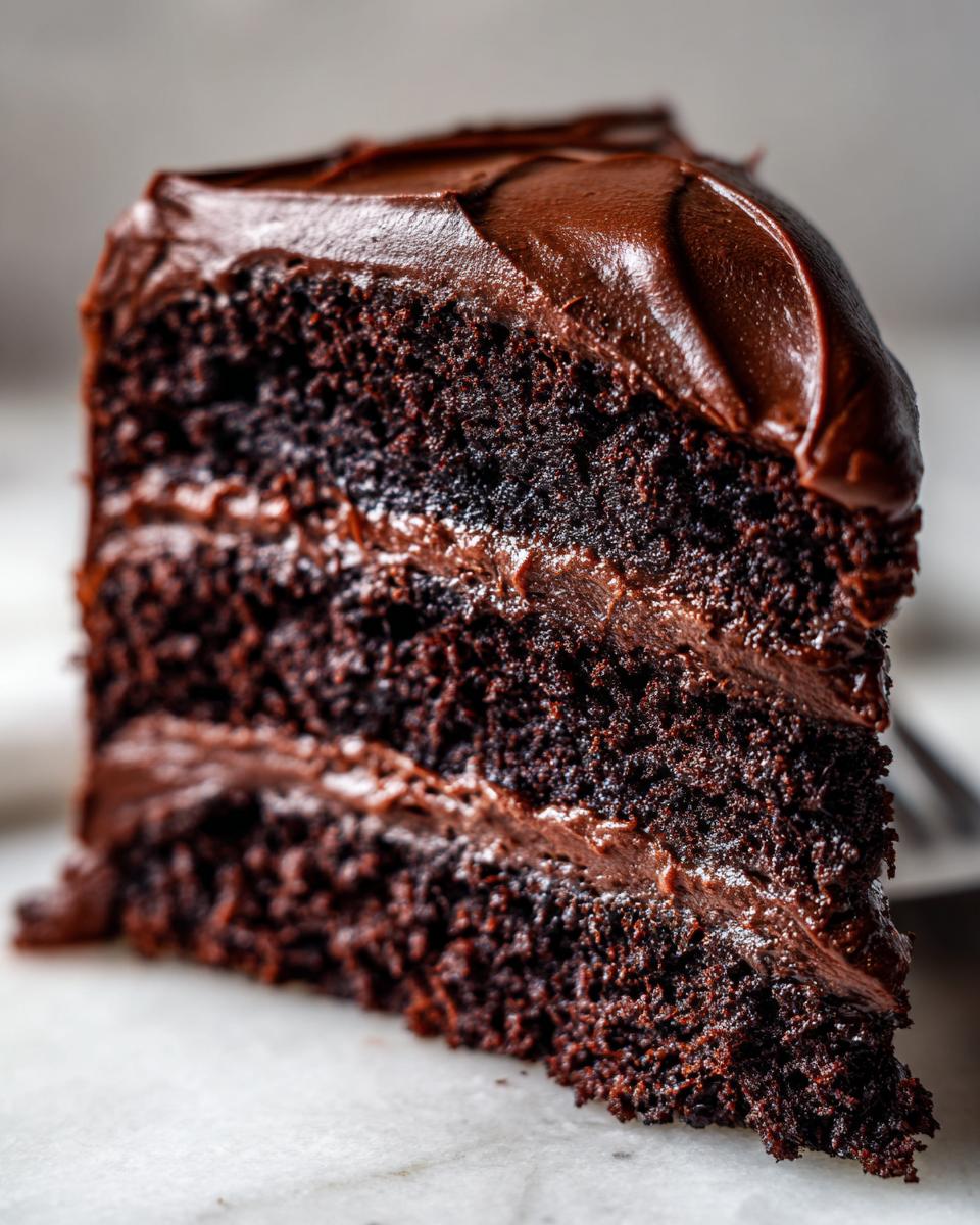 Close-up of a decadent slice from the chocolate cake recipe, showing three moist layers and thick chocolate frosting.