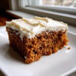 A close-up of a moist slice of spice cake topped with thick cream cheese frosting, sitting on a white plate.