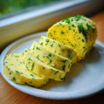 A log of bright yellow garlic butter mixed with green herbs, sliced on a white plate.