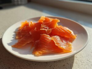 Close-up of bright orange, flaky slices of smoked salmon piled on a small white plate, illuminated by sunlight.