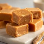 Close-up of rich, caramel-colored penuche fudge cut into perfect squares stacked on a white marble surface.