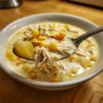 A spoonful of creamy chicken corn chowder featuring shredded chicken, corn, and potato chunks being lifted from a white bowl.