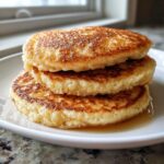 Close-up of a stack of three golden-brown, fluffy pancakes soaking in a small pool of syrup on a white plate.