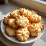 A pile of golden, star-shaped butter cookies stacked on a white ridged plate, illuminated by sunlight.