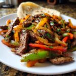 Close-up of seasoned steak fajitas mixed with colorful bell peppers and onions, served with a tortilla.