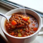 A spoonful of steaming, rich lentil soup filled with lentils, carrots, and celery being lifted from a white mug.