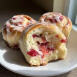 Three soft Strawberry Cheesecake Sweet Rolls on a white plate, one is broken open revealing strawberry filling and cream cheese.