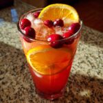 A tall glass filled with bright red thanksgiving punch, ice, fresh cranberries, and orange slices.