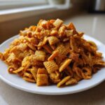 A mound of crunchy, glossy toffee chex mix recipe piled high on a white plate, ready to eat.