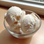 Two scoops of creamy vanilla protein ice cream served in a small clear glass bowl on a wooden surface.