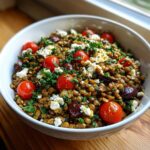 Close-up of a hearty lentil salad topped with bright red cherry tomatoes, feta cheese crumbles, olives, and fresh parsley.