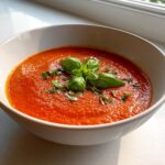 Close-up of a creamy, bright red bowl of tomato basil soup garnished with fresh basil leaves.