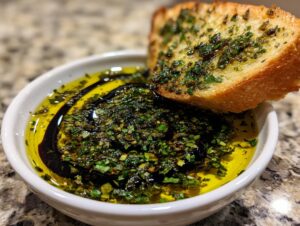 Close-up of a small white bowl containing olive oil bread dip with herbs and balsamic vinegar, served with a piece of toasted bread.