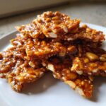 Close-up of broken, glossy pieces of homemade peanut brittle recipe stacked on a white plate.