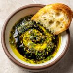 Close-up of a crusty piece of bread being dipped into an olive oil bread dip mixture with balsamic vinegar and herbs.