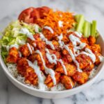 A close-up of a vibrant buffalo chicken bowl featuring saucy chicken pieces over white rice, drizzled with ranch, and topped with fresh vegetables.