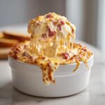 A spoonful of hot, cheesy reuben dip is lifted from a white bowl, showing long, gooey cheese pulls.