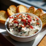 A white bowl filled with creamy bruschetta dip topped with sun-dried tomatoes and herbs, served with toasted baguette slices.