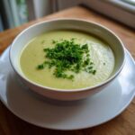 A close-up of a bowl of creamy celery soup, topped generously with chopped fresh parsley.