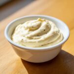 A close-up of thick, creamy homemade garlic aioli swirled in a small white bowl on a wooden surface.