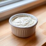 A small white ramekin filled with thick, creamy horseradish sauce speckled with spices, sitting on a wooden surface.