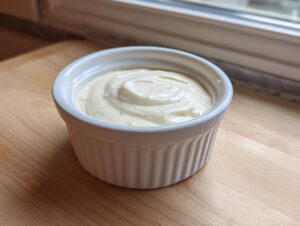 A small white ramekin filled with creamy, pale yellow horseradish sauce, sitting on a light wooden surface near a window.