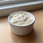 A white fluted ramekin filled with creamy, freshly whipped horseradish sauce, sitting on a wooden surface near a window.