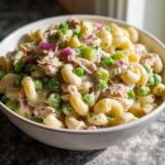 A close-up, appetizing view of creamy Tuna Pasta Salad featuring elbow macaroni, peas, red onion, and celery in a white bowl.