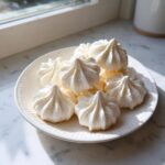A close-up of several crisp, white meringue cookies piped into swirls, resting on a speckled white plate.